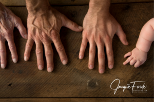 Three hands in a row on a wooden surface: an elderly person's hand, an adult's hand, and a baby's hand.