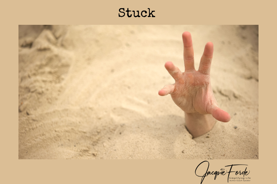 A hand emerging from sand, with fingers spread wide, against a beige background. The word "Stuck" is written at the top. Jacquie Forde's logo and slogan are in the bottom right corner.