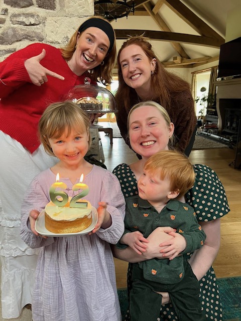 A young girl holds a birthday cake with "62" candles, surrounded by two women, a woman holding a toddler, and one woman pointing and smiling. They are indoors in a cozy living room.